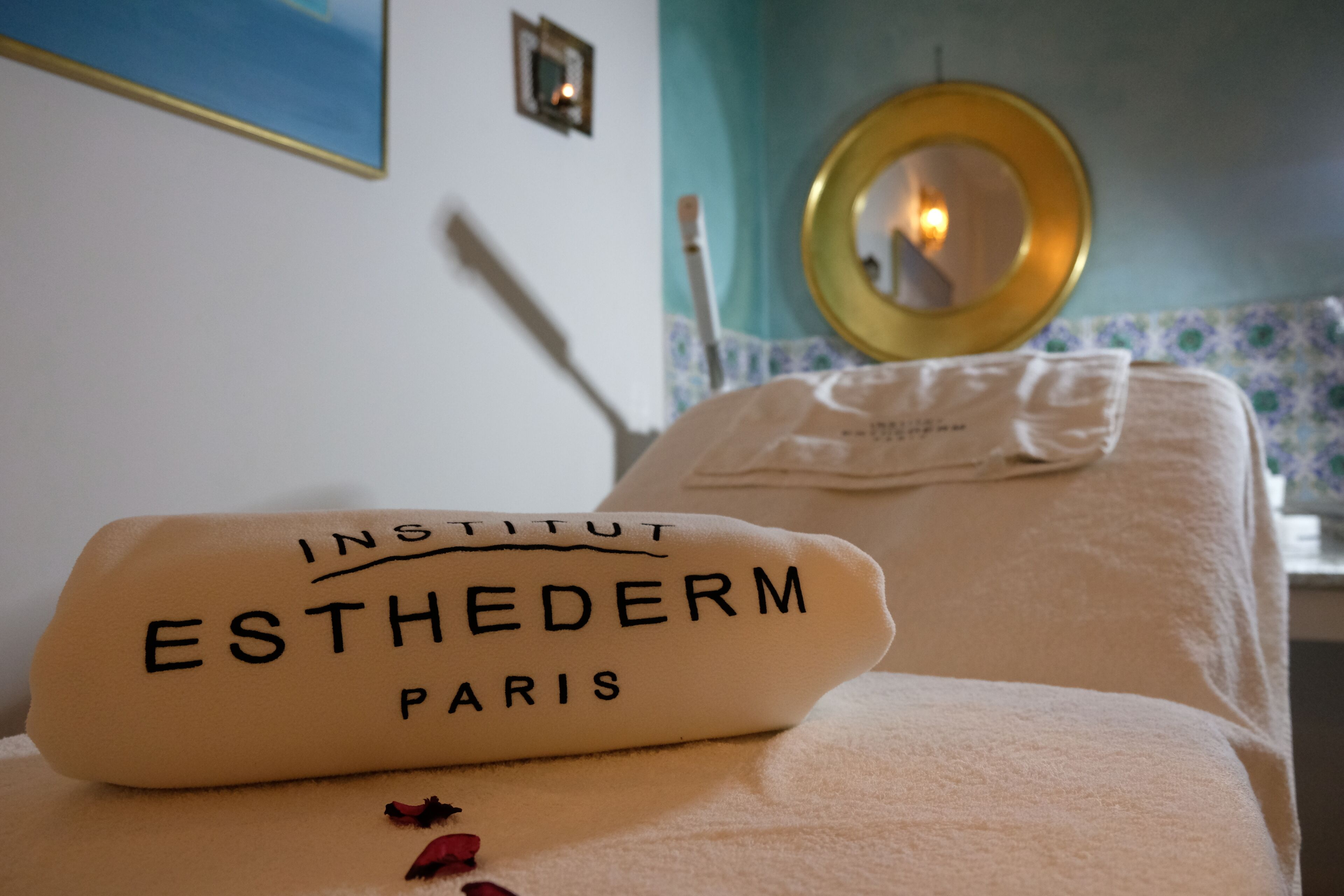 Una habitación de hotel con una cama, una toalla con el texto 'INSTITUT ESTHEDERM PARIS' y un espejo en la pared.