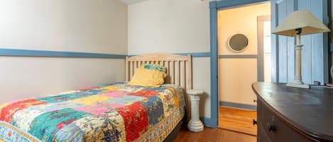 Un dormitorio con una cama de madera, un colcha colorida, una mesita de noche blanca pequeña, un aparador y una puerta que da a otra habitación.