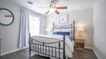 Ein Schlafzimmer mit einem Bett, einem Deckenventilator, einem Nachttisch mit Lampe und einem Fenster mit Vorhängen.