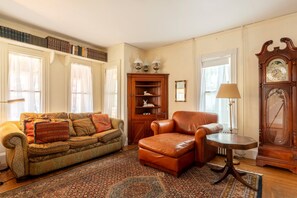 A living room with a sofa, armchair, a wooden cabinet with books, a clock, and a lamp. - Full Lovely Home Walking Distance to Town & Harbor (Mount Desert)