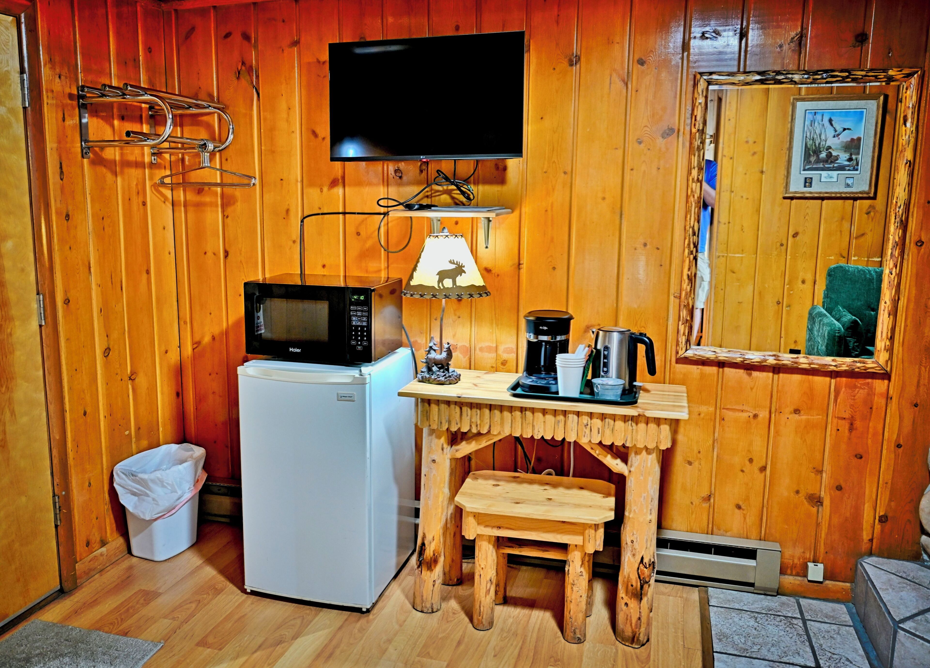 Habitación pequeña con detalles de madera, un refrigerador, microondas, cafetera, una mesa con lámpara y un espejo.