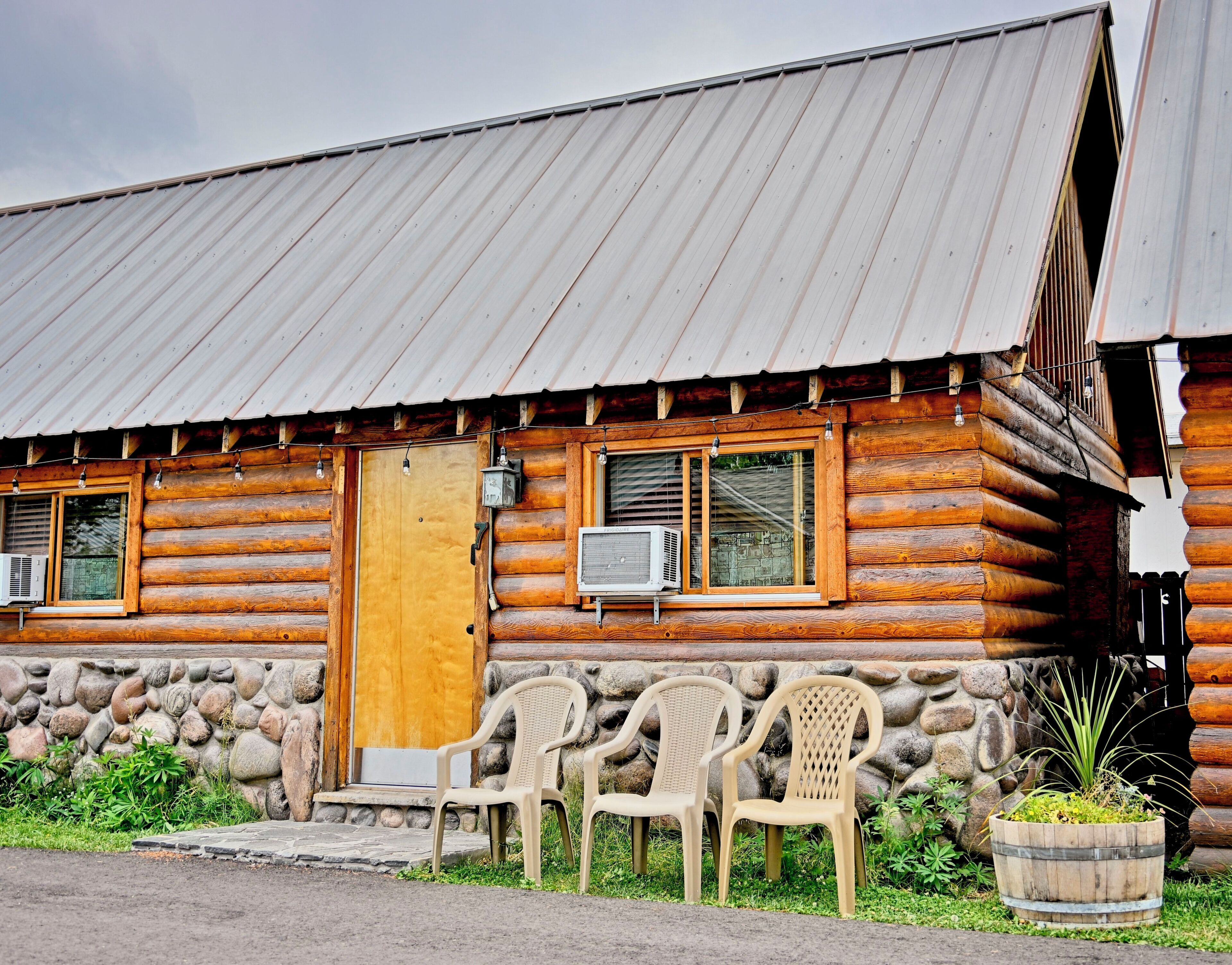 A log cabin with a metal roof, a wooden door, and a window with an air conditioning unit. There are plastic chairs outside and a stone wall with a planter.