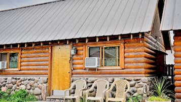 Une cabane en rondins avec un toit en métal, une porte en bois et une fenêtre équipée d’un appareil de climatisation. À l’extérieur, on trouve des chaises en plastique et un mur de pierres agrémenté d’un jardinière.