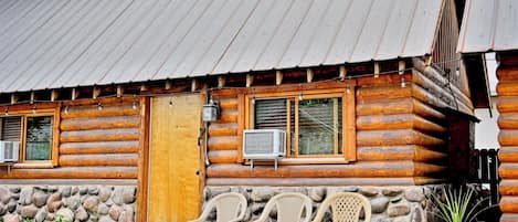 A log cabin with a metal roof, a wooden door, and a window with an air conditioning unit. There are plastic chairs outside and a stone wall with a planter.