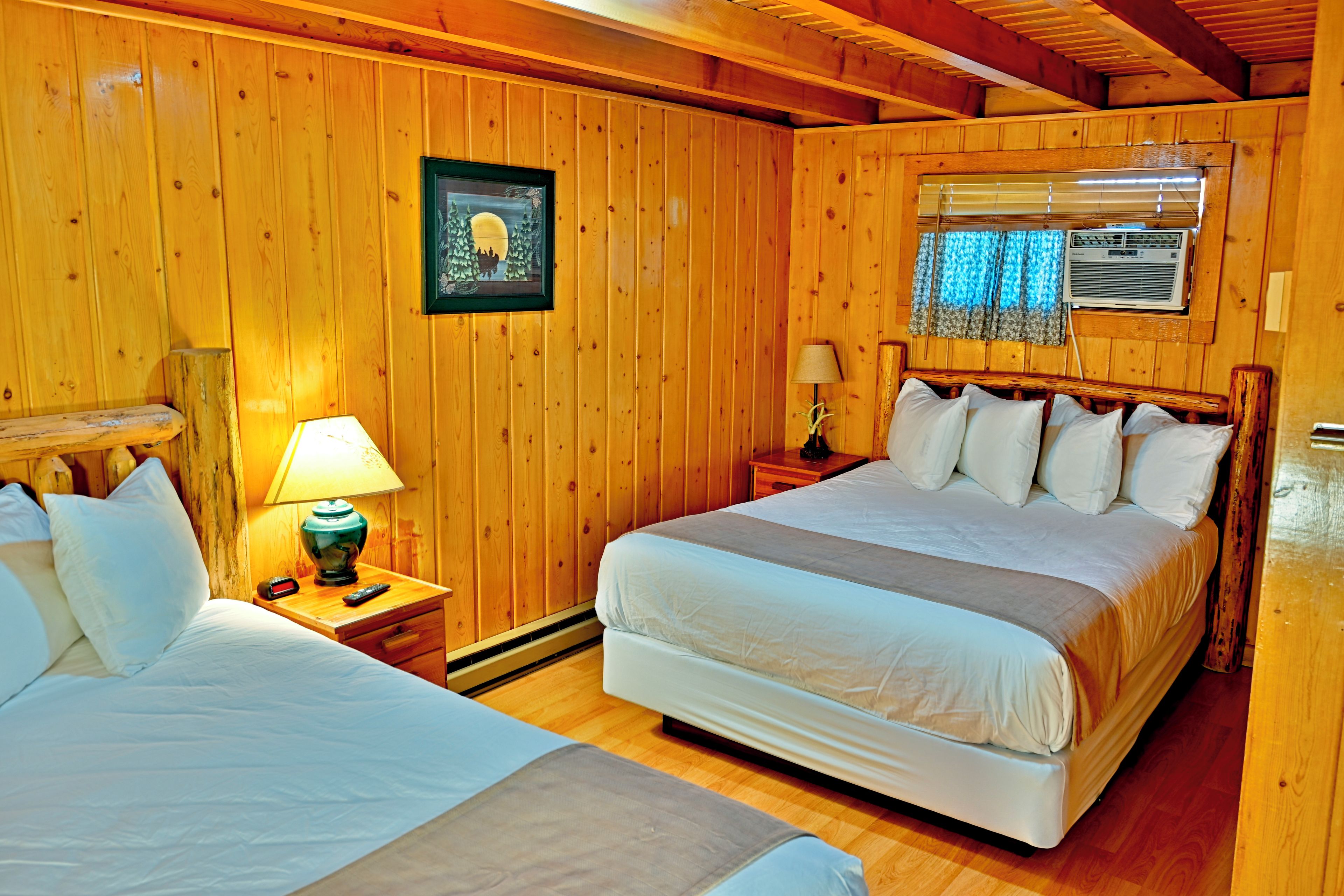 Una habitación con paneles de madera, dos camas, una mesita de noche con una lámpara, un cuadro en la pared y una ventana con persianas.