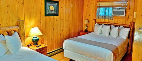 A wooden-paneled room with two beds, a nightstand with a lamp, a framed picture on the wall, and a window with blinds.