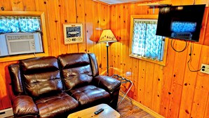 A room with a leather sofa, a small wooden table, a lamp, a television, and a window with curtains.