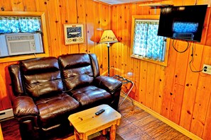 A room with a leather sofa, a small wooden table, a lamp, a television, and a window with curtains.