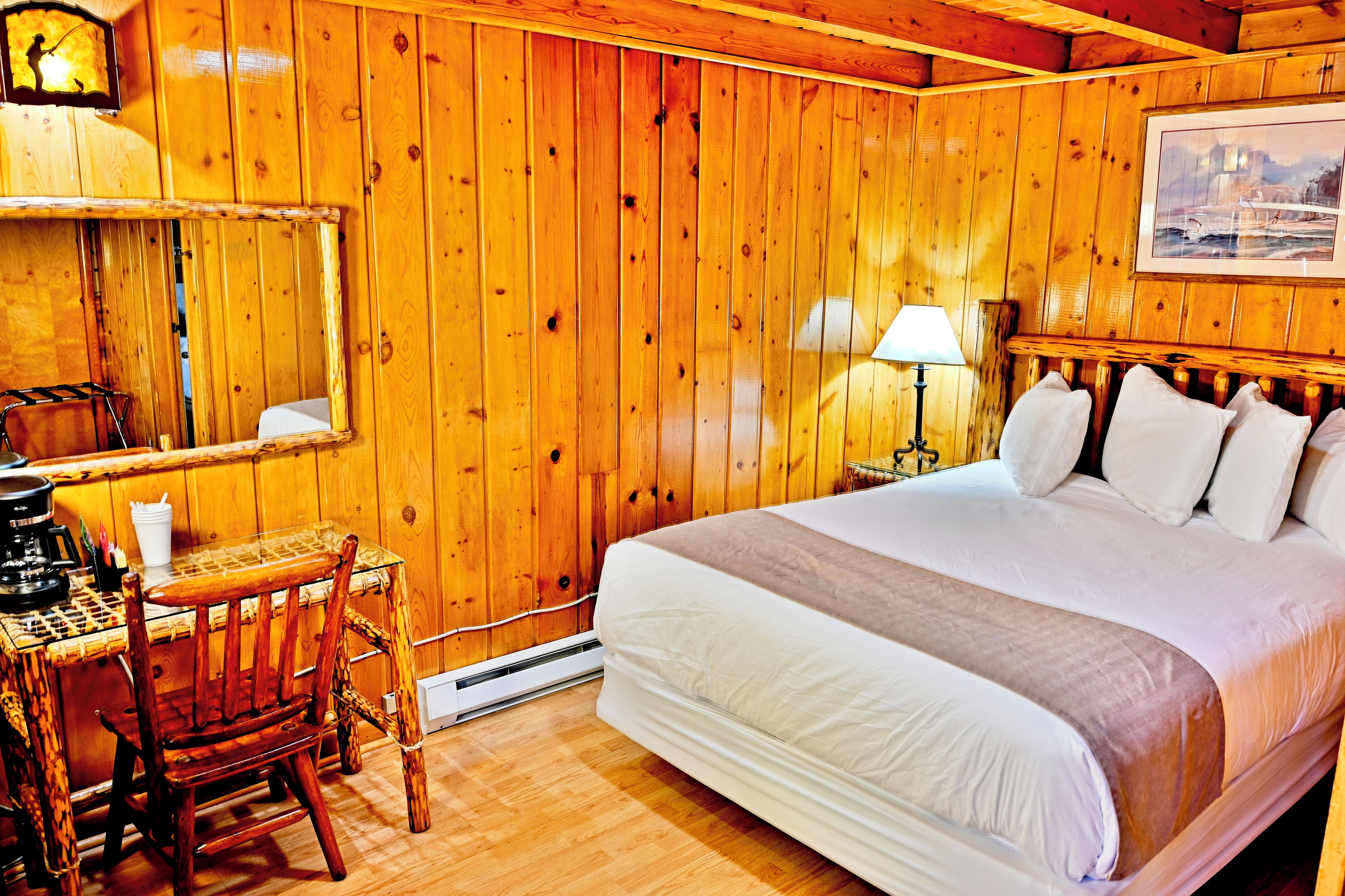 Una habitación de madera con una cama, un escritorio, una silla, una lámpara y un cuadro en la pared.