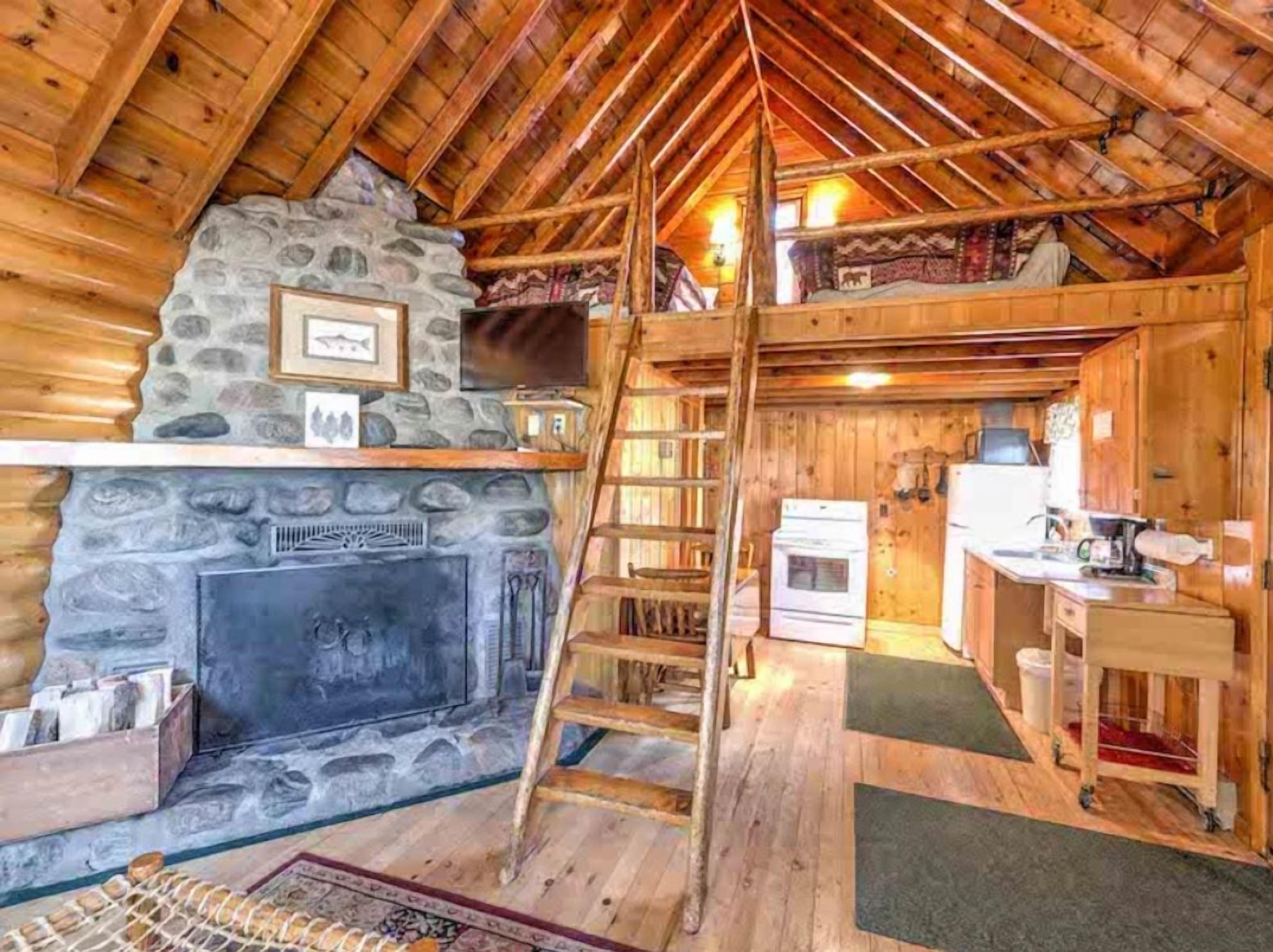 A log cabin interior with a stone fireplace, wooden beams, a ladder, a kitchen area, and a loft space.