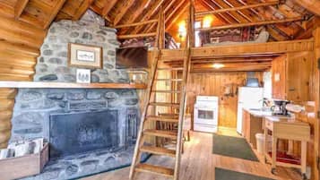 A log cabin interior with a stone fireplace, wooden beams, a ladder, a kitchen area, and a loft space.