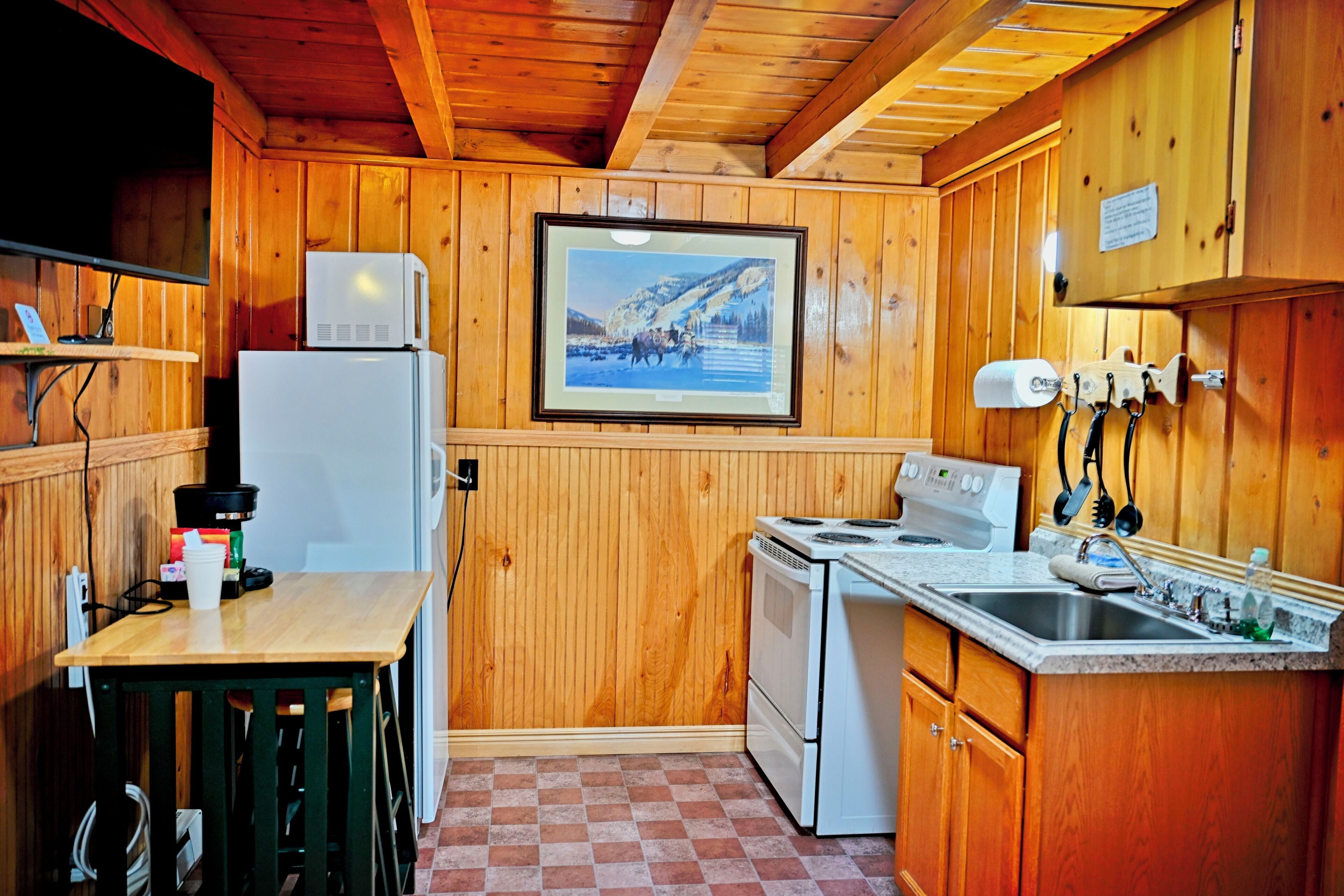 A small kitchen with wooden walls, a refrigerator, a stove, a microwave, a sink, and a painting on the wall.
