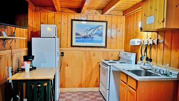A small kitchen with wooden walls, a refrigerator, a stove, a microwave, a sink, and a painting on the wall.