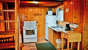A compact kitchen with wooden walls, a stove, a refrigerator, a sink, and a coffee maker.