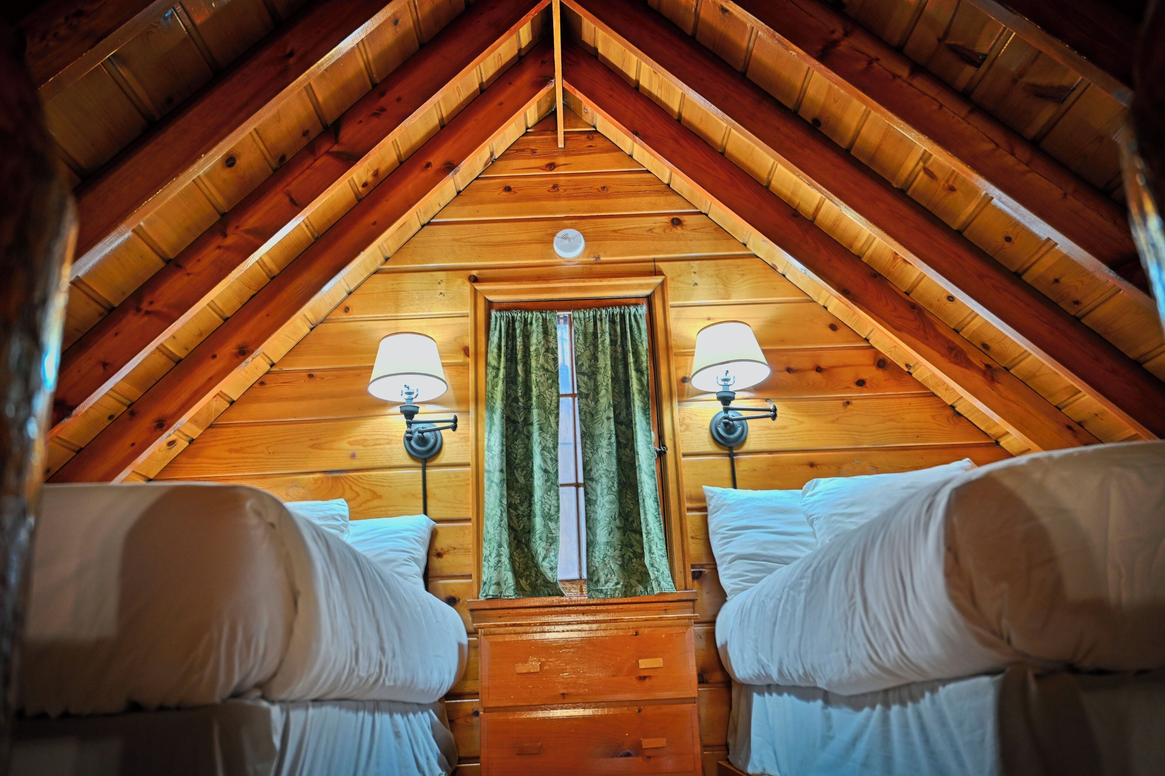 Ein gemütliches Schlafzimmer im Dachgeschoss mit schrägverdeckter Holzbalkendecke, zwei Nachttischlampen, einem fenster mit Vorhängen und einem Bett mit weißen Leinen.