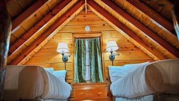 Ein gemütliches Schlafzimmer im Dachgeschoss mit schrägverdeckter Holzbalkendecke, zwei Nachttischlampen, einem fenster mit Vorhängen und einem Bett mit weißen Leinen.