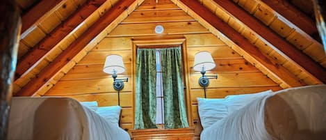 A cozy attic bedroom with a sloped wooden ceiling, two bedside lamps, a window with curtains, and a bed with white linens.