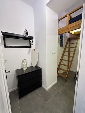 Une pièce avec une commode noire, une étagère fixée au mur ornée d’un objet décoratif, une échelle et une porte donnant sur une autre pièce.