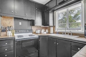 Une cuisine avec des meubles de rangement de couleur sombre, une cuisinière, un évier et une fenêtre avec des stores.