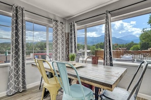 A dining area with a table and chairs, large windows with curtains, and a view of mountains and greenery. - Pet-Friendly 2BR Penthouse with Mountain Views + Private Deck (Colorado Springs)