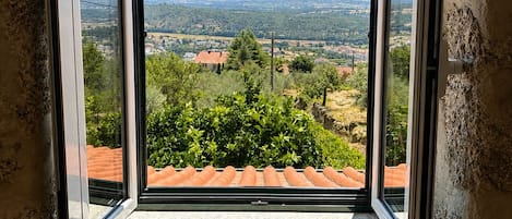A room with a large window offering a view of a rural landscape with trees and distant buildings.