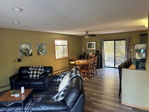 A living room with a sofa, coffee table, and a dining area with a table and chairs. There is a ceiling fan, a window with blinds, and a water dispenser. - Casa de la Playa - Walk to the beach (Rimbey)