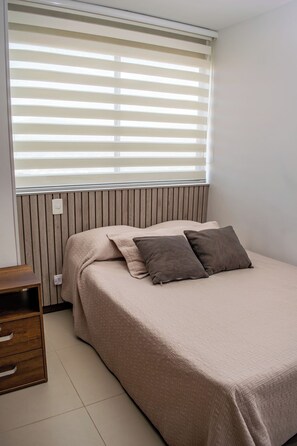 A bedroom with a bed, a wooden nightstand, and a window with blinds. - Luxury 3BR Panoramic Views – El Tesoro Medellín (Medellín)