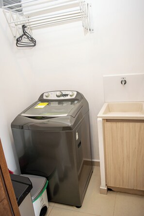 A compact laundry room with a front-loading washing machine, a drying rack, and a small sink. - Luxury 3BR Panoramic Views – El Tesoro Medellín (Medellín)