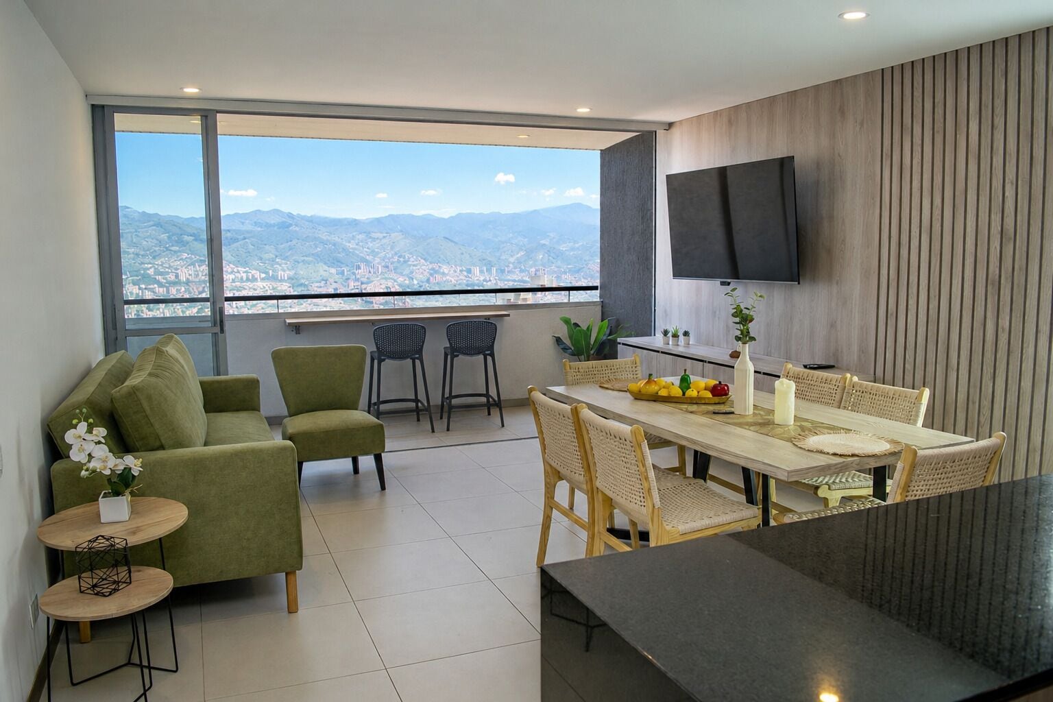 A modern living room with a sofa, chairs, a dining table with a fruit bowl, and a TV mounted on the wall.