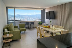 A modern living room with a sofa, chairs, a dining table with a fruit bowl, and a TV mounted on the wall. - Luxury 3BR Panoramic Views – El Tesoro Medellín (Medellín)