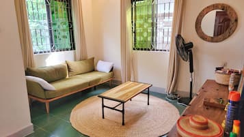 A room with a sofa, a round woven rug, a wooden table, a standing fan, and a mirror on the wall.