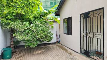 A paved courtyard with a metal gate, a green trash bin, and lush green foliage.