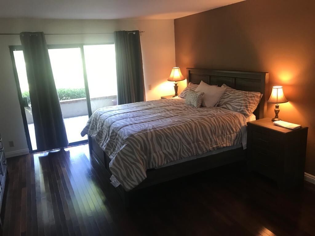 A bedroom with a bed, two bedside tables with lamps, a window with curtains, and a view of the outdoors.