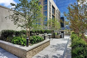 Un patio al aire libre con mobiliario, plantas y un edificio moderno al fondo.