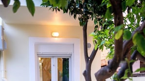 Uma área de pátio com porta de vidro que dá para um espaço interior, mobiliário de exterior branco e uma árvore em vaso.