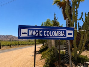 A directional sign for 'MAGIC COLOMBIA' with a right-pointing arrow, set against a backdrop of a clear sky, a dirt road, and desert vegetation.