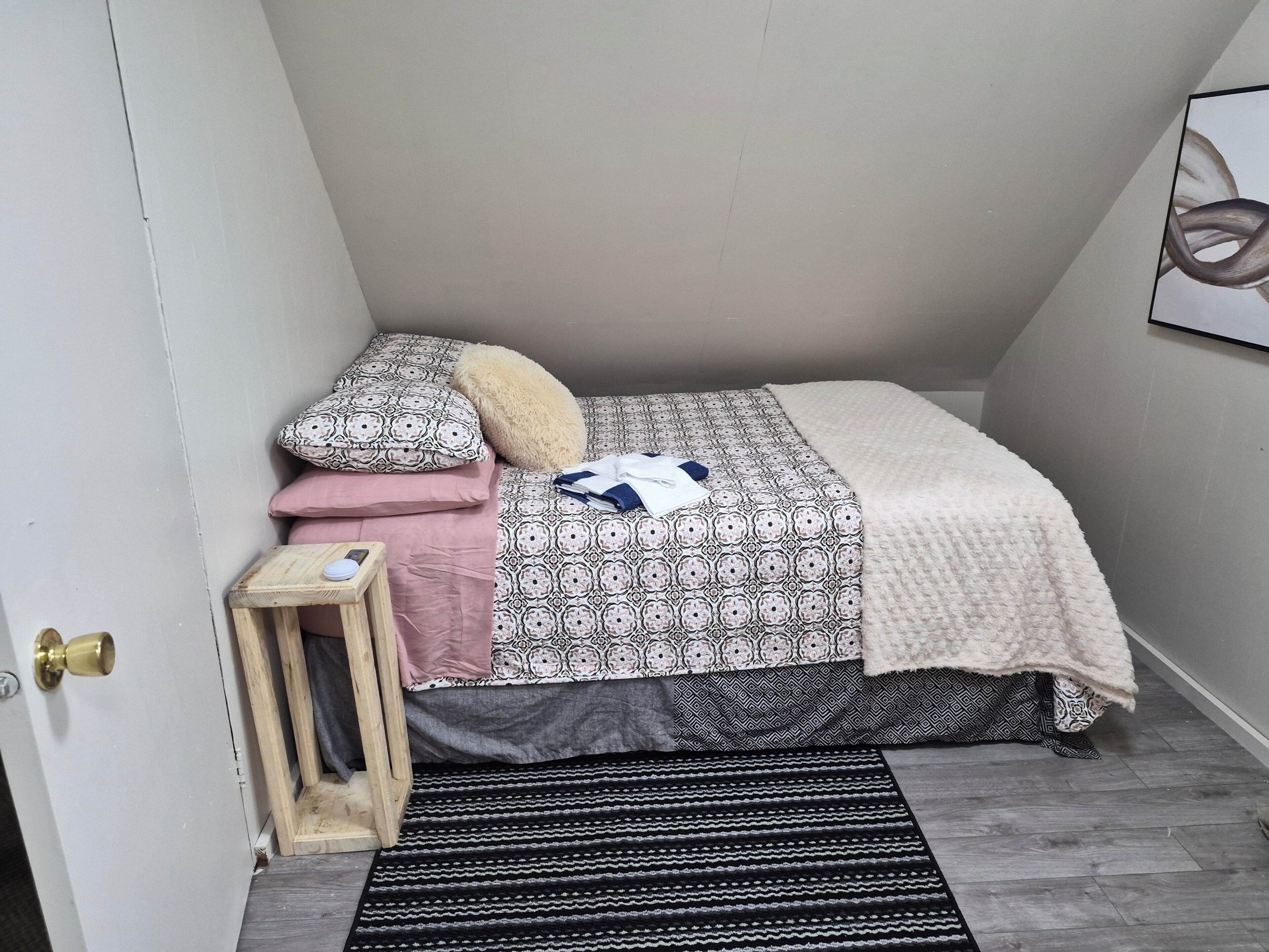 A bedroom with a bed, pillows, a blanket, a small wooden stool, and a black and white striped rug.