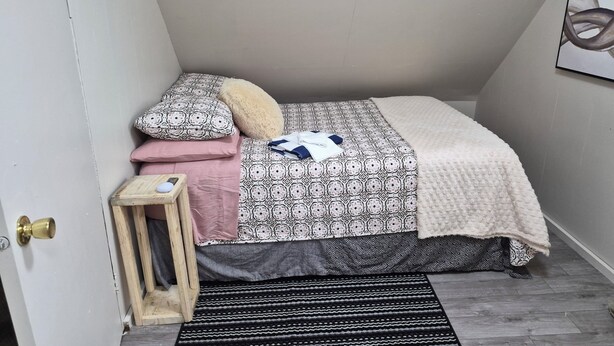 A bedroom with a bed, pillows, a blanket, a small wooden stool, and a black and white striped rug.