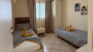 A bedroom with two beds, a bedside table, a wardrobe, and a window with curtains. - HostySpot - Tropical Resort, 2-Bedroom Apartment (Santa Maria)