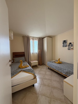 A bedroom with two beds, a bedside table, a wardrobe, and a window with curtains. - HostySpot - Tropical Resort, 2-Bedroom Apartment (Santa Maria)