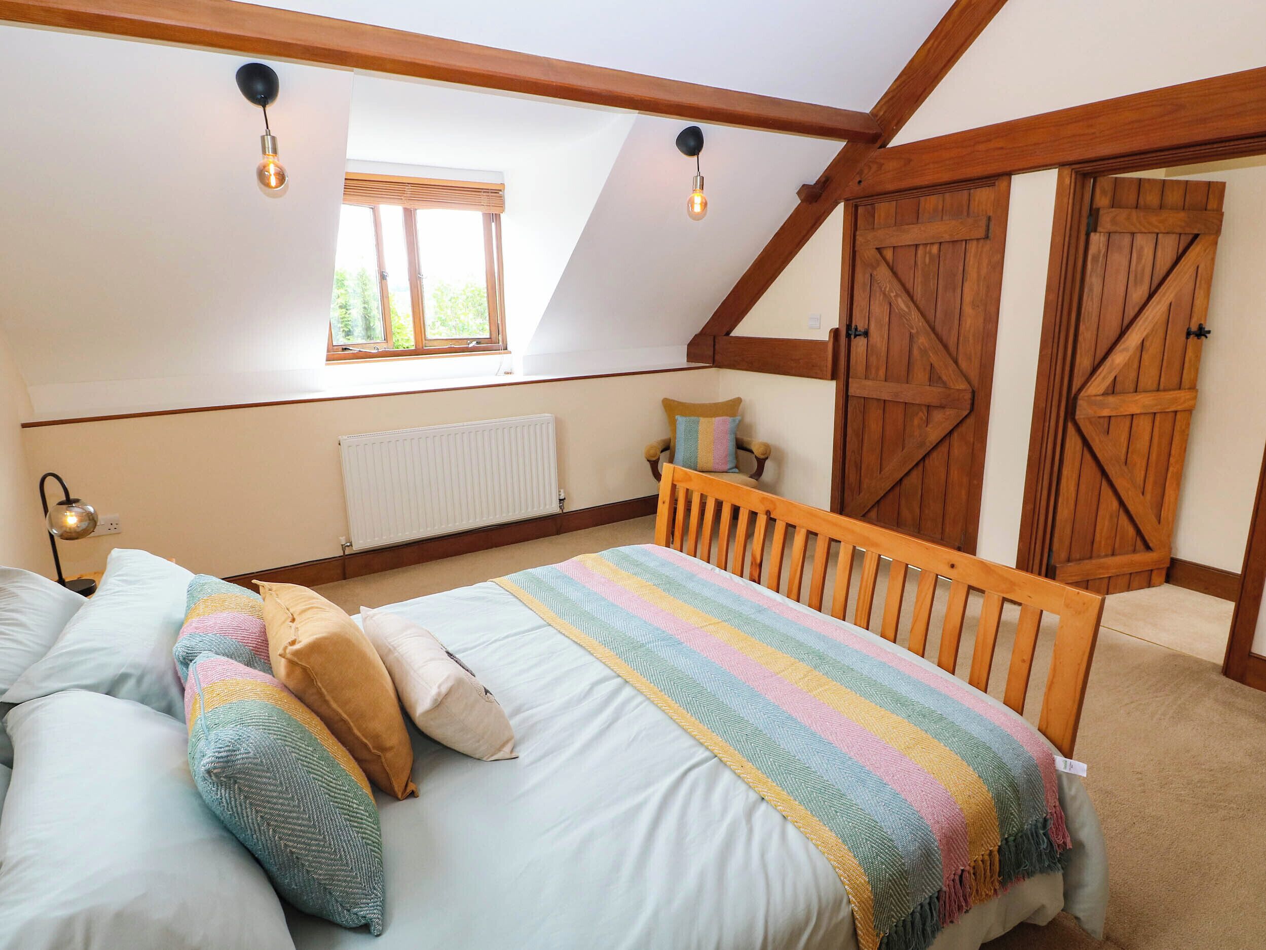 Ein Schlafzimmer mit schrägen Wänden, einem Bett mit gestreifter Decke, Holzbalken, einer Heizung, einem Fenster und einer Tür.