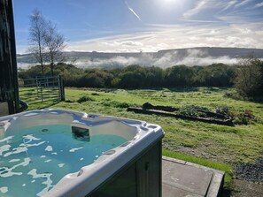 A hot tub with a view of a grassy field and a forested hill in the background. - Penrhiwoli Barn (Llangammarch Wells)