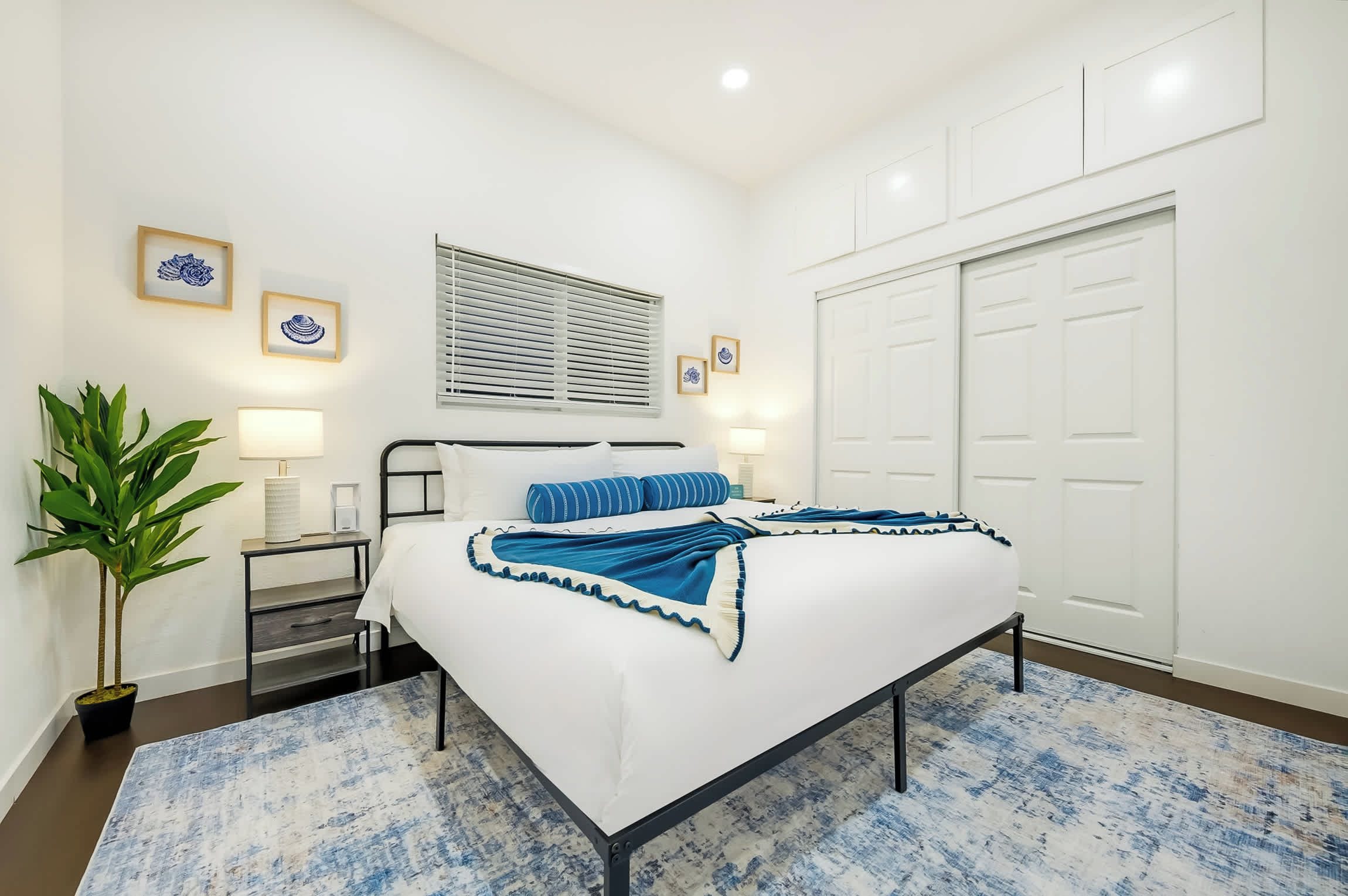 Une chambre à coucher bien rangée, avec un lit, des tables de chevet, une plante en pot et une fenêtre avec des stores.