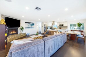 A modern living room with a large sectional sofa, a fireplace, and a kitchen area with white cabinets and stainless steel appliances. - Pacific Grand - Dual Living Near Mission Bay (San Diego)