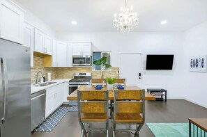 A modern kitchen with white cabinets, stainless steel appliances, a wooden dining table with chairs, and a chandelier. - Pacific Grand - Dual Living Near Mission Bay (San Diego)