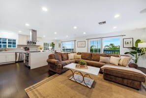 A modern living space with a kitchen, a large brown sectional sofa, and a coffee table with a plant. - Pacific Grand - Dual Living Near Mission Bay (San Diego)