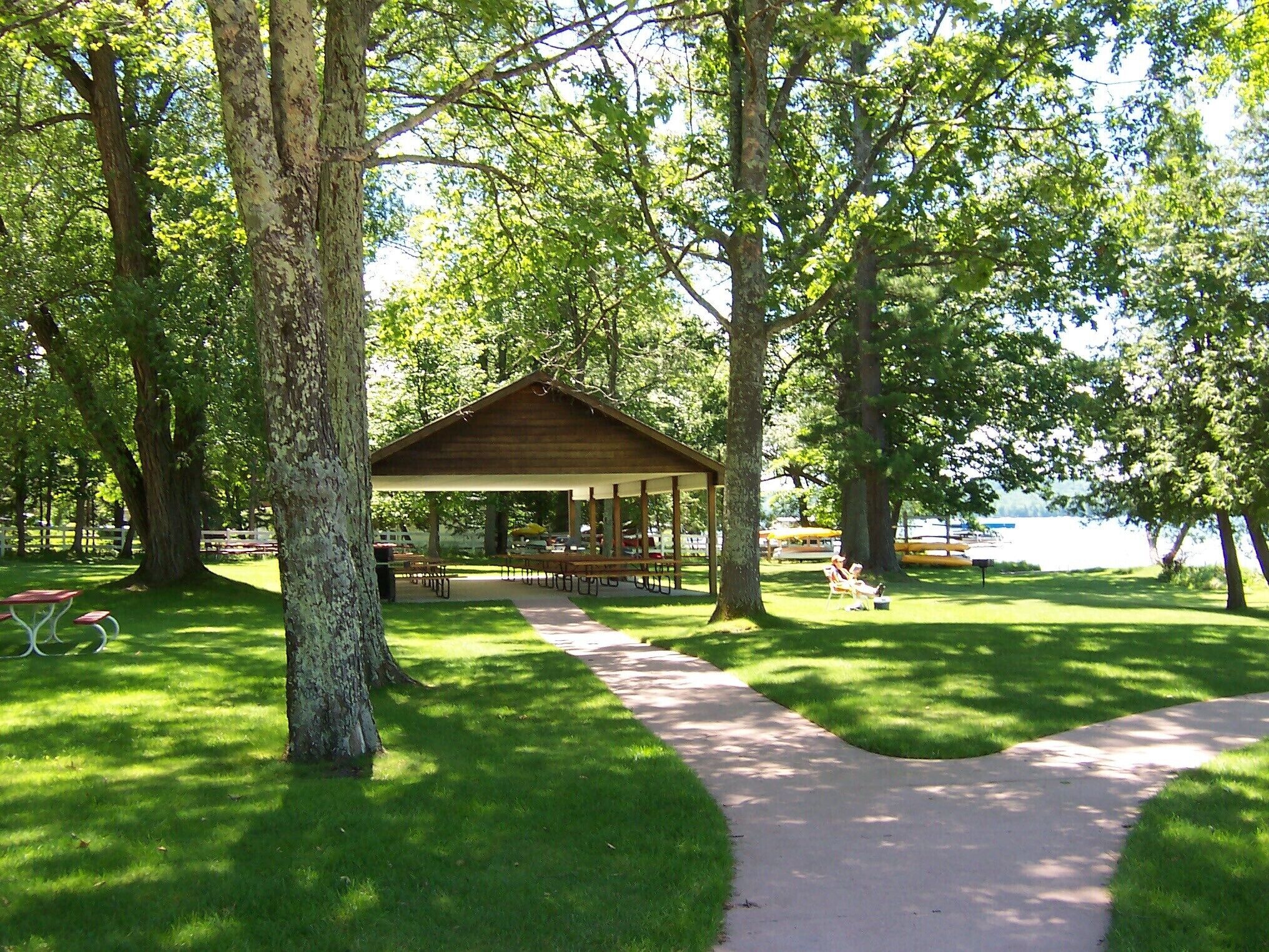 A covered picnic area with benches and tables, surrounded by trees and a pathway leading to a lake.