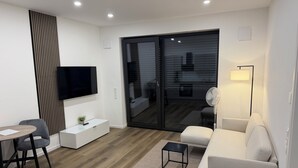 Een moderne woonkamer met een flatscreen televisie, een bank, een salontafel, een eethoek met stoelen en een glazen schuifdeur naar een buitengebied.