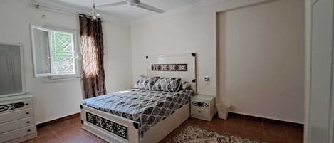 A bedroom with a bed, a nightstand, a ceiling fan, a window with curtains, and a patterned rug.