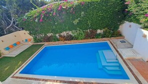 Una piscina rectangular amb superfície d'aigua blava i cristal·lina, envoltada per una paret blanca, una tanca de vegetació i plantes amb flors.